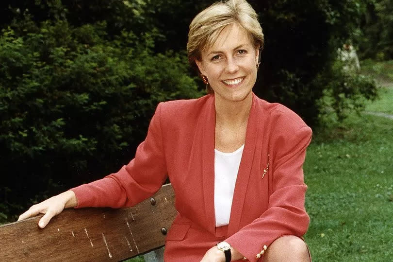 Jill Dando in a red suit jacket and skirt sat on a park bench