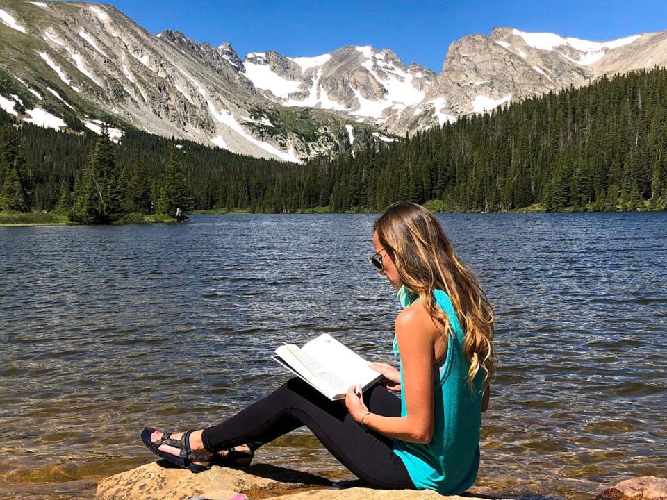 Emily sits on a rock near a lake and reads a book. The lake is surrounded by trees and the mountains in the background have a dusting of snow.