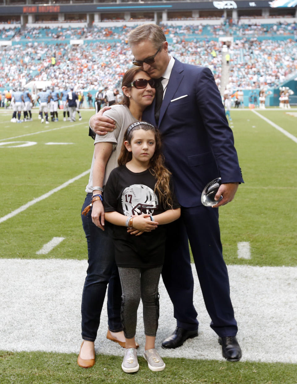 FILE - Tom Garfinkel, Miami Dolphins President and CEO, hugs Melissa Feis during a presentation honoring her husband, Marjorie Stoneman Douglas High School Football Coach Aaron Feis, during the second half of an NFL football game against the Tennessee Titans, Sunday, Sept. 9, 2018, in Miami Gardens, Fla. At the center is Feis' daughter Arielle. Aaron Feis was honored as the Miami Dolphins 2018 George F. Smith Coach of the Year. (AP Photo/Wilfredo Lee, File)