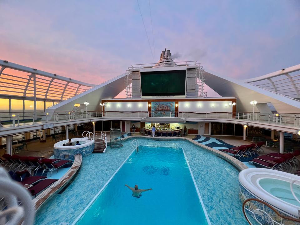 Pool on top of a cruise ship during sunset with a screen playing movies above the pool