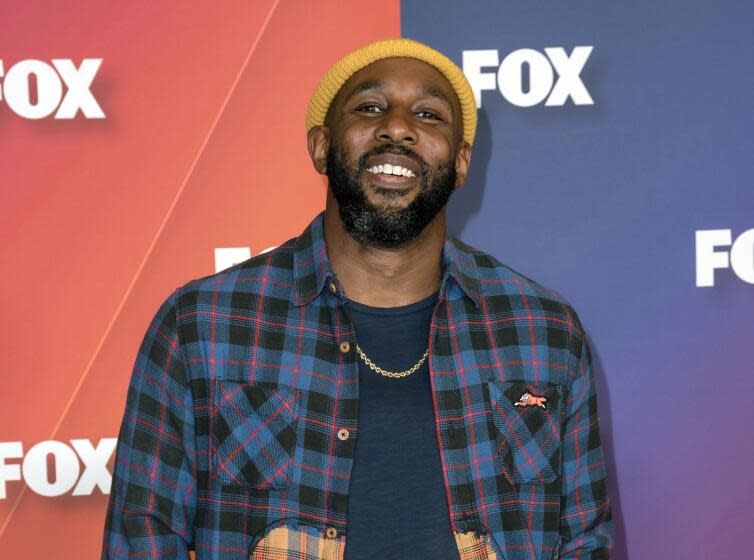 A man in a yellow beanie, blue flannel shirt and gold chain tilting his head up and smiling