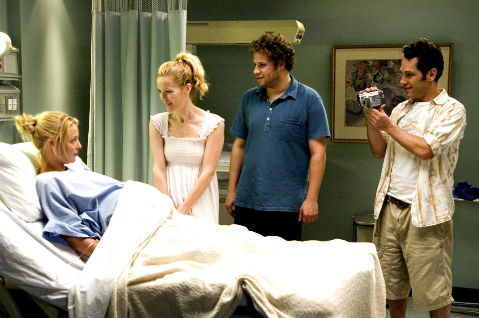 Katherine Heigl is in a hospital bed with Leslie Mann, Seth Rogen, and Paul Rudd standing beside her. Paul Rudd is holding a camcorder