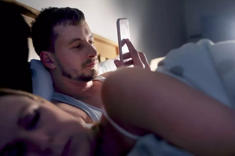 Man looking at phone in bed with woman sleeping beside him