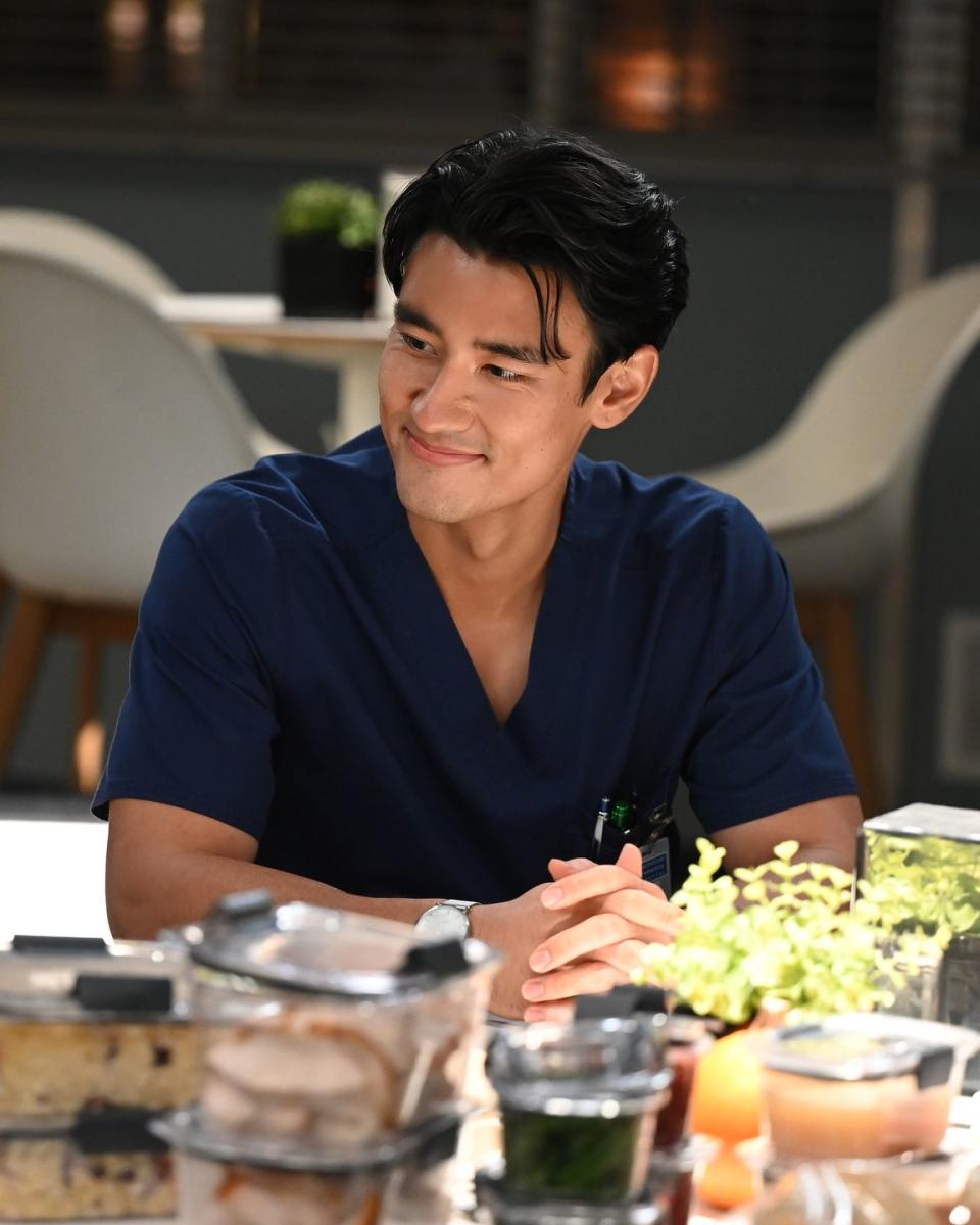 actor alex landi as dr nico kim in season 18 of grey's anatomy, shown in wearing a navy scrub shirt and smiling at someone off camera