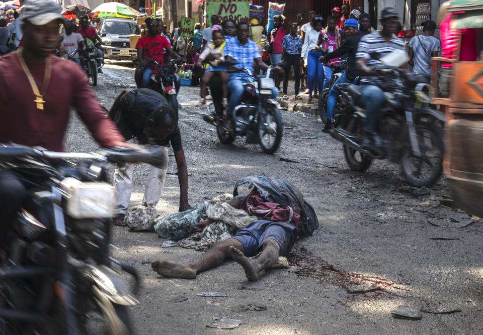 FILE - A body lies in the street as commuters, including a little girl on the back of a motorcycle, top center, make their way through the Petion-Ville neighborhood of Port-au-Prince, Haiti, April 22, 2024. As young Haitians are increasingly exposed to violence, the country is undergoing a wider push to dispel a long-standing taboo on seeking therapy and talking about mental health. (AP Photo/Ramon Espinosa, File)