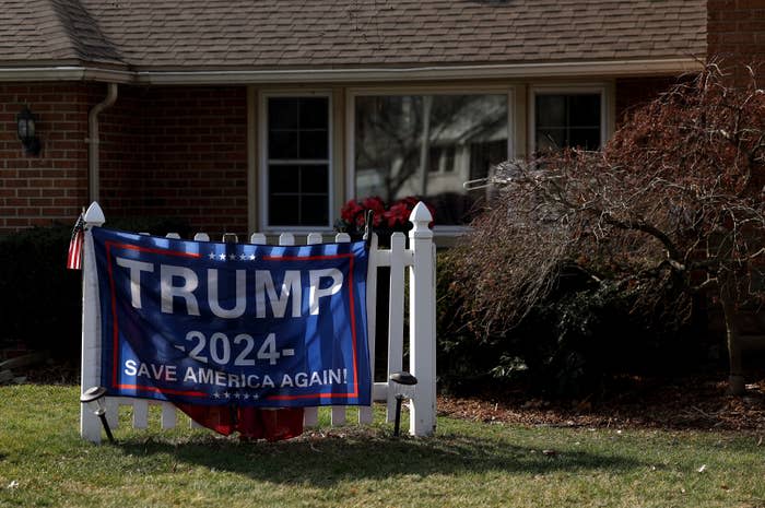 A white fence in front of a house displays a "Trump 2024 - Save America Again!" banner