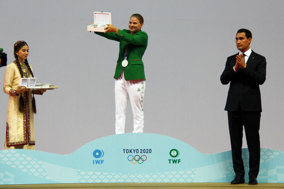 Polina Guryeva, Olympic silver medalist in the women's 59kg weightlifting event, at the 2020 Summer Olympics, center, shows her Presidential gift as Turkmenistan's authoritarian president, Gurbanguly Berdymukhamedov's son Serdar, who was recently promoted to deputy prime minister, applauds her at a lavish ceremony in Ashgabat, Turkmenistan, Saturday, Aug. 21, 2021. Turkmenistan has lauded its first Olympic medal winner at a lavish ceremony where she was showered with gifts. Guryeva, 21, lifted a total 217 kilograms in the 59-kilogram category, edging Mikiko Andoh of Japan for second place at the Tokyo Games. (AP Photo/Alexander Vershinin)