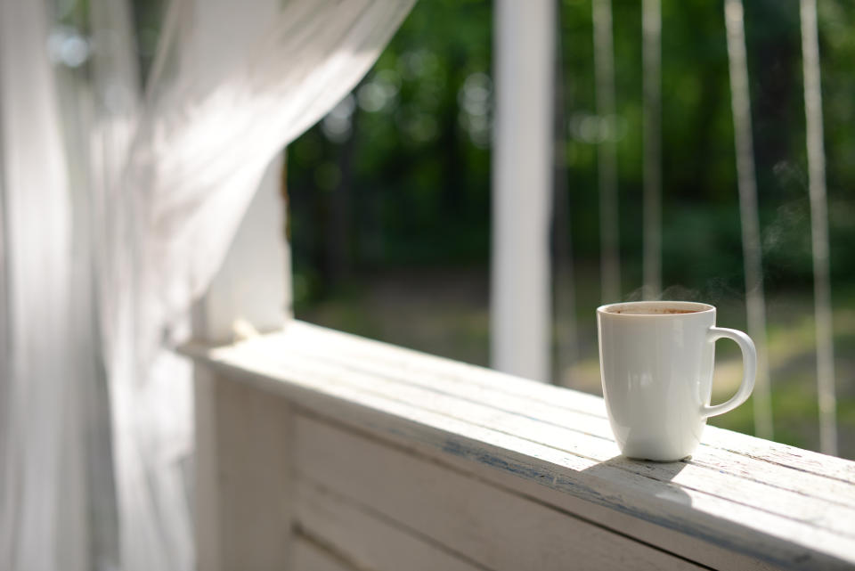 A white mug of coffee sits on a wooden porch railing with flowing drapes and a background of greenery