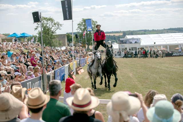 A crowd watches a man standing the backs of two horses