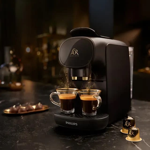 L'OR Barista Sublime coffee machine in black on a marble countertop next to two coffee pods and a plate of chocolate.