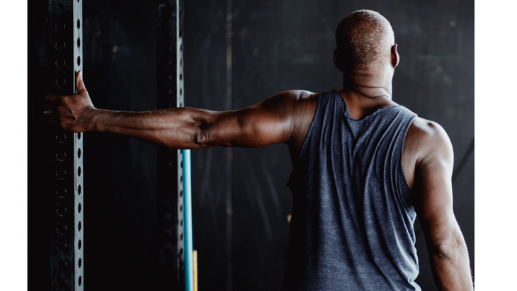 Man in a tank top at the gym stretching his shoulder by placing one hand on a bar