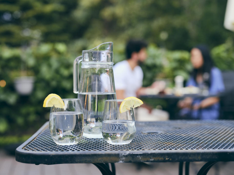 Water pitcher and glasses with lemon slices on an outdoor table, blurry background of two people chatting in a garden setting