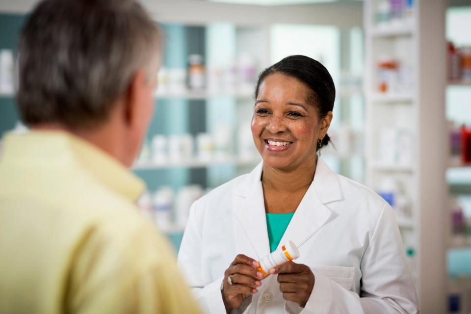 A smiling pharmacist holding a prescription bottle in their hands while speaking with a customer. 