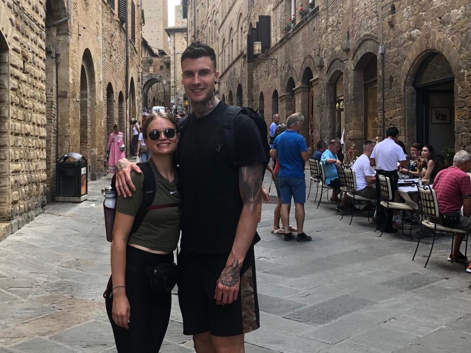 Photo of Emily and Jakob standing close together and smiling.   Jakob has his right hand on Emily's shoulder and they are standing on a stone path with restaurant tables filled with people. Beyond that are brown brick buildings. Emily wears sunglasses, green t-shirt, black leggings, and black sneakers. Jakob wears a black t-shirt, black shorts, and black sneakers. His tattoos on his left hand and bicep are visible.