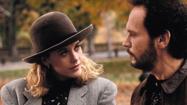 meg ryan in bowler hat and black and white striped jacket with padded shoulders looking at billy crystal in beard and brown leather jacket they are sitting outside in fall and looking serious