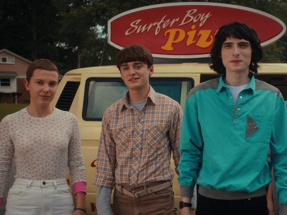 eleven, will, mike, jonathan, and argyle in stranger things, standing in front of a yellow van with a sign on top that says surfer boy pizza