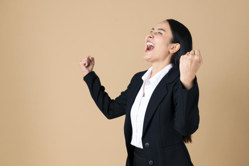 Studio shot of cheering businesswoman with arms raised into fists, illustrating a story about asking for promotions at work.