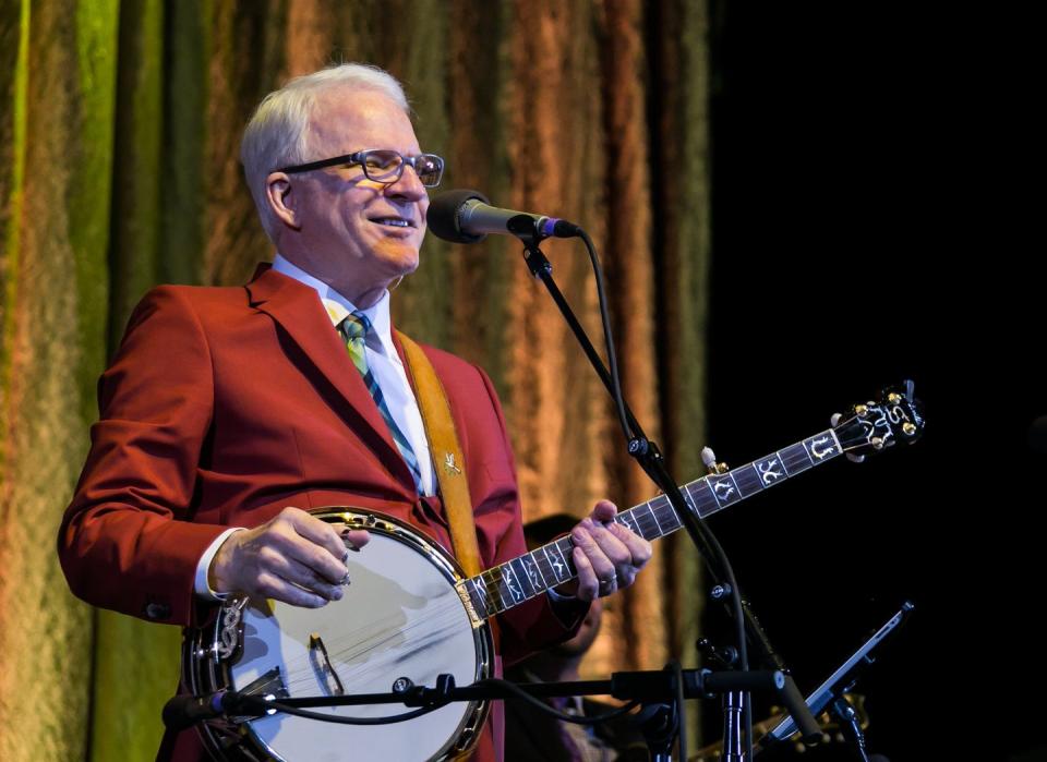 steve martin smiles while standing behind a microphone and holding a banjo