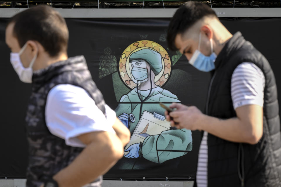 People walk by a depiction of a medical staff wearing protective equipment, executed in the style of orthodox icons, in Bucharest, Romania, Wednesday, April 29, 2020. The artwork, among others depicting medical staff in the manner of religious icons, created by designer Wanda Hutira, is part of a campaign called Thank You Doctors, meant to raise awareness to the work of medical staff fighting the COVID-19 pandemic. Following public pressure by Romania's influential Orthodox church the artworks, described as "blasphemous" will be removed from all locations in the Romanian capital, according to the agency behind the project. (AP Photo/Andreea Alexandru)