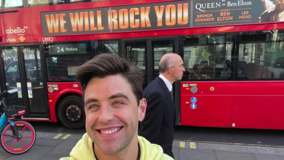 Jarryd Nurden in a yellow top posing in front of a London bus with a We Will Rock You
