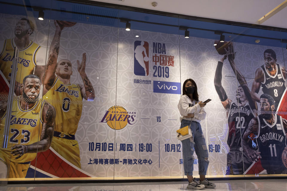 In this Friday, Oct. 11, 2019, photo, a woman wearing a mask stands near promotion boards for a NBA preseason game between Brooklyn Nets and Los Angeles Lakers in Beijing. When Houston Rocket's general manager Daryl Morey tweeted last week in support of anti-government protests in Hong Kong, everything changed for NBA fans in China. A new chant flooded Chinese sports forums: "I can live without basketball, but I can't live without my motherland."(AP Photo/Ng Han Guan)