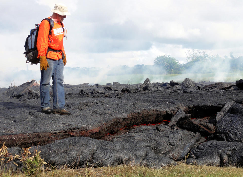 This Oct. 25, 2014 photo provided by the U.S. Geological Survey shows a Hawaii Volcano Observatory geologist standing on a partly cooled section of lava flow near the town of Pahoa on the Big Island of Hawaii. Note the thin red horizontal line of molten lava visible along the bottom third of the photo. The flow here is about one meter (three feet) thick, but slightly farther upslope where the lava has had more time to inflate the thickness was closer to two meters. Dozens of residents in this rural area of Hawaii were placed on alert as flowing lava continued to advance. Authorities on Sunday, Oct. 26, 2014 said lava had advanced about 250 yards since Saturday morning and was moving at the rate of about 10 to 15 yards an hour, consistent with its advancement in recent days. The flow front passed through a predominantly Buddhist cemetery, covering grave sites in the mostly rural region of Puna, and was roughly a half-mile from Pahoa Village Road, the main street of Pahoa. (AP Photo/U.S. Geological Survey)