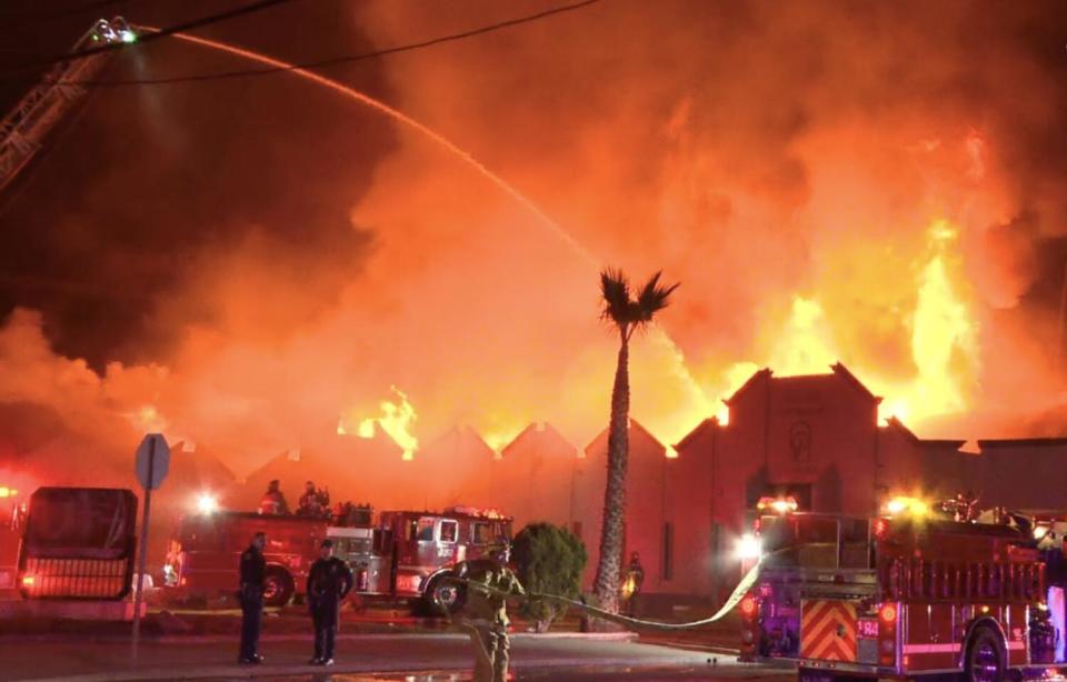 Firefighters and a firetruck outside a large building that is engulfed in flames.