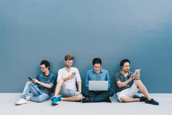 Multiethnic group of four men using smartphone, laptop computer, digital tablet together with copy space on blue wall