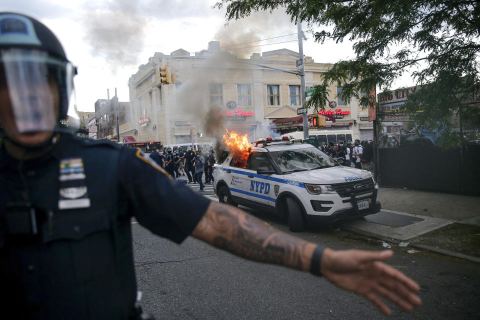 Police officers in riot gear push back protesters as a police car with NYPD painted on the side burns during a demonstration.