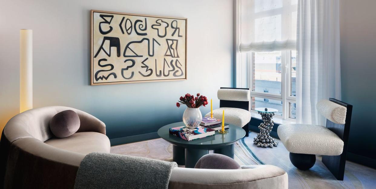 living room with ombre painted walls in shades from palest white blue to darker blue at the base along with a horizontal piece of art with black hieroglyphic type characters on it and a curved sofa in a off gray fabric and two armless chairs with column base and a matte blue round table at center