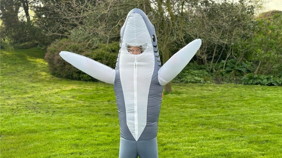 Georgie Box in her shark fancy dress. Georgie is a 28-year-old white woman and is pictured inside an inflated shark costume with both arms held up. The shark's smiling mouth provides a window through which you can just about make out Georgie's blonde hair. She's pictured outside in a park on a bright day