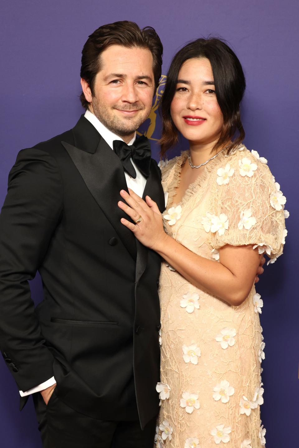 Michael Angarano in a black tuxedo and Maya Erskine in a floral dress on a purple carpet