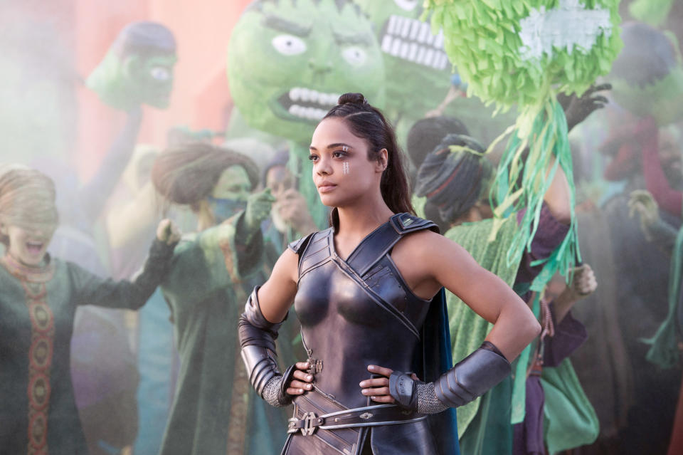 Tessa Thompson as Valkyrie stands confidently in armored costume amidst a vibrant festival with people holding up giant green Hulk masks in the background