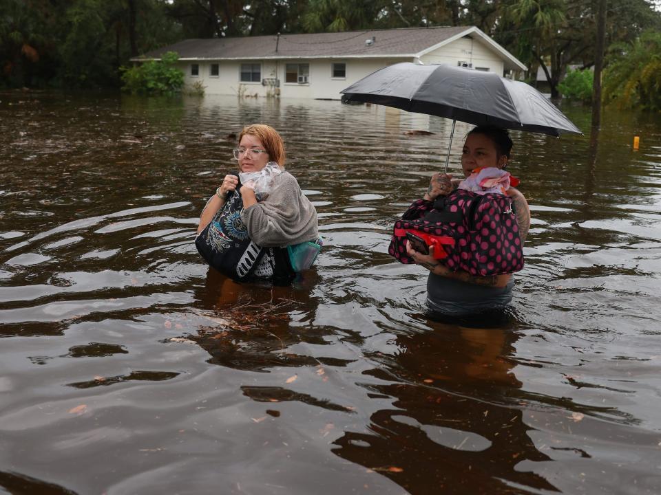 two women carrying bags and an umbrella wade through waist-high brown flood waters with a flooded house in the background