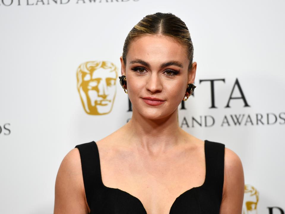 Sophie Skelton wearing a black dress and large hoop earrings while standing in front of a sign for the Baftas.