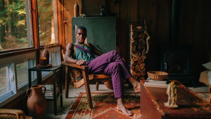 A man with purple pants sits in a dark living room full of shadow and carpets