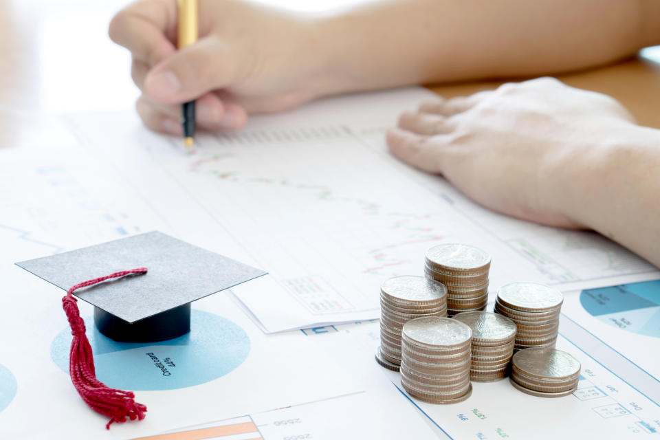 person writing on a piece of paper with a small graduation cap and stacks of coins in the foreground