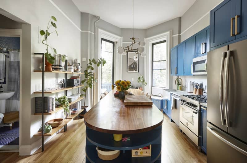 The kitchen is painted gray, has blue cabinetry, open shelves filled with plants and stainless steel appliances.