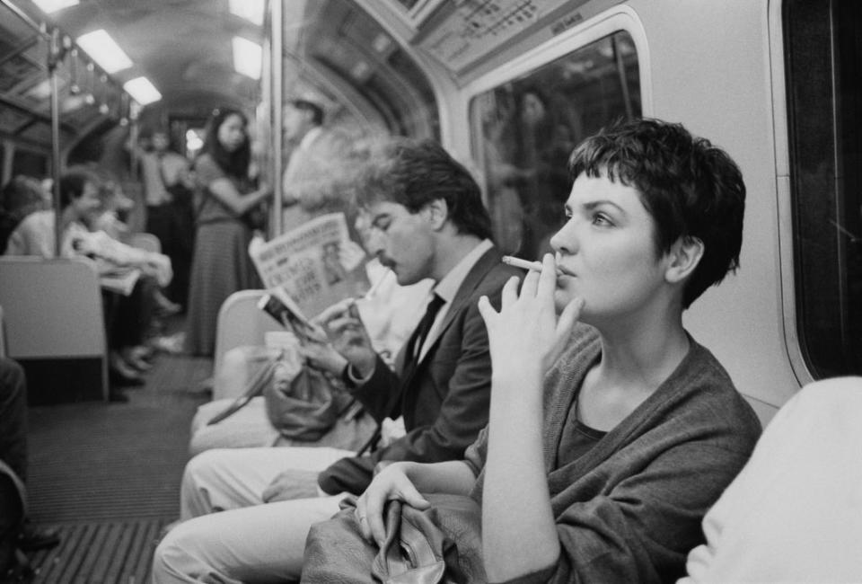 Woman smoking next to a man reading a newspaper on a subway, other passengers in background