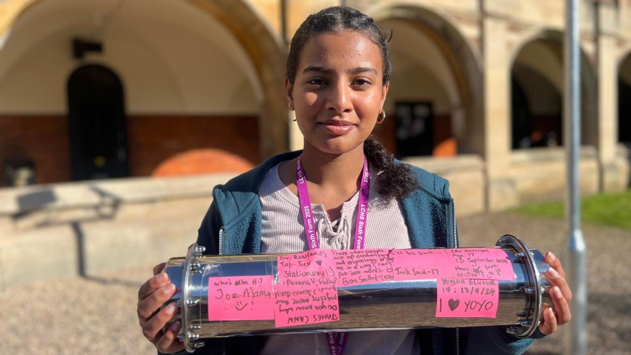 A girl with dark hair, tied back into a ponytail, wears a blue zip-up hoodie and stripe top. She smiles at the camera, standing in front of the school, while holding the time capsule. The time capsule is covered in pink coloured messages created by the students.