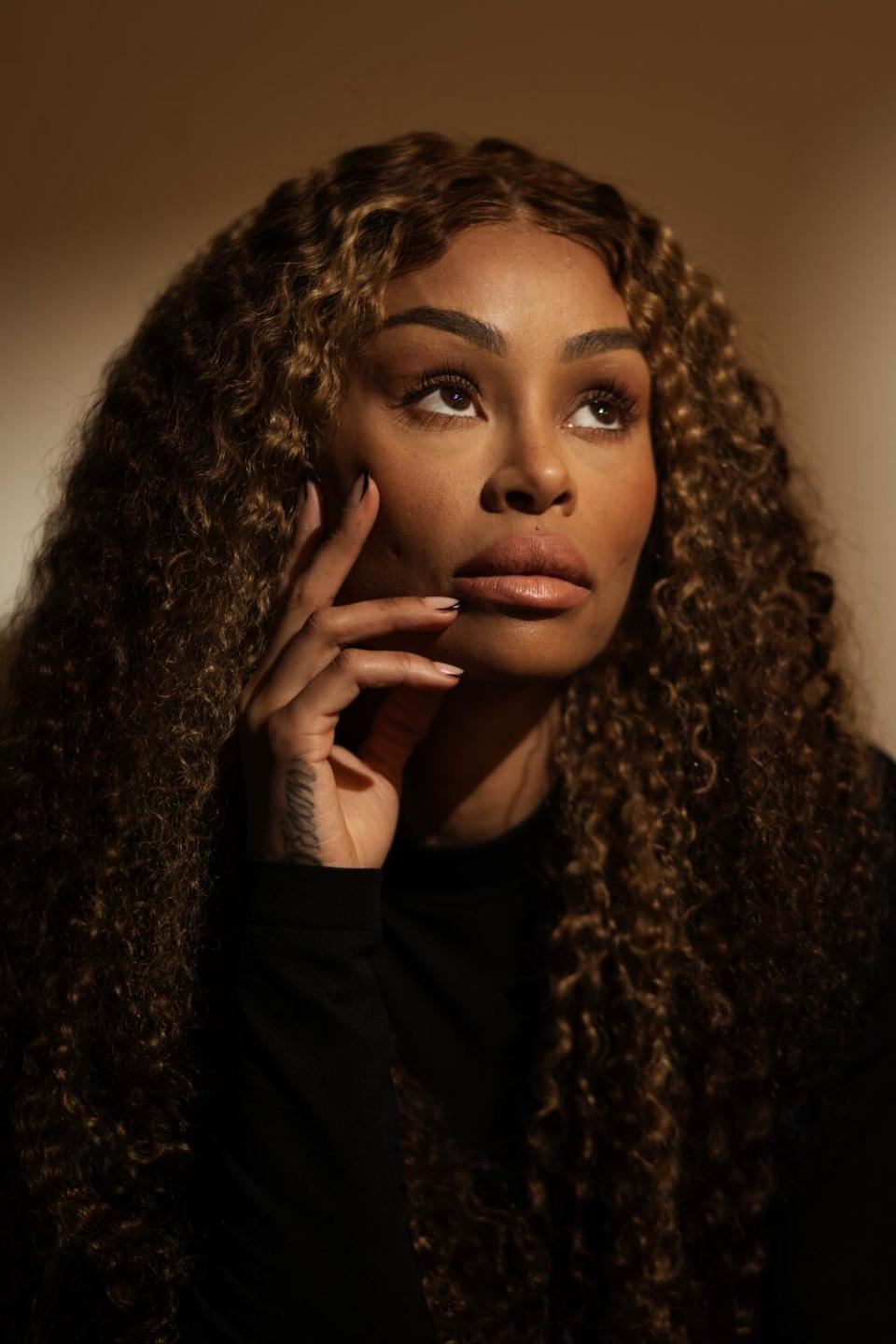 A woman with long hair holds her hand against her face and looks pensively to the right.