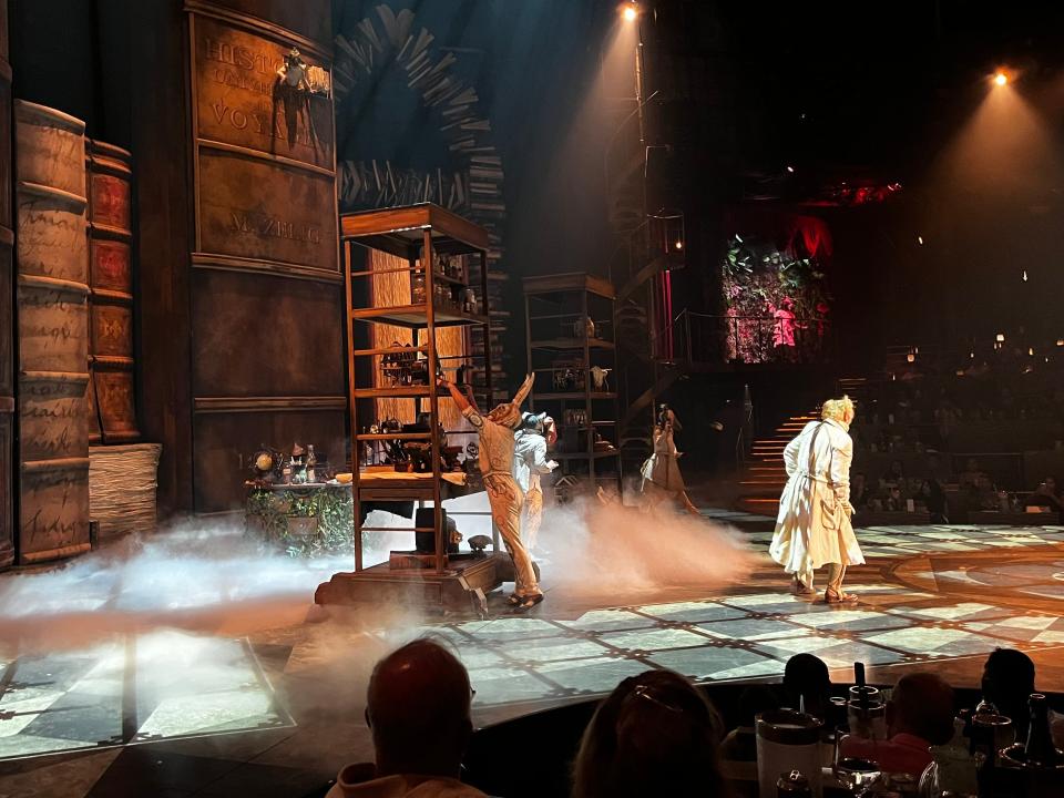 Cirque du Soileil performers dressed in white dresses and other clothing wearing masks. A character wearing a rabbit mask unloads items from a tall wooden cart on the stage. Smoke surrounds the characters