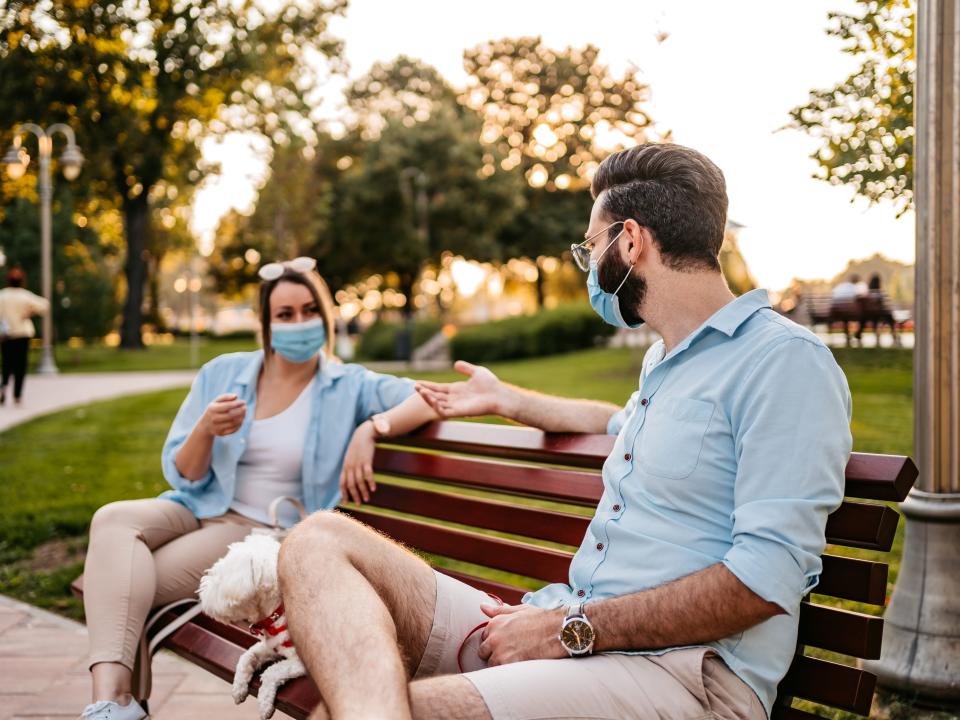 Young cute heterosexual couple holding social distance on park bench and wearing protective face masks. Small dog sitting between them.