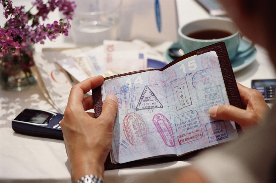 Hands holding an open passport filled with various stamps on a table with a mobile phone and tea