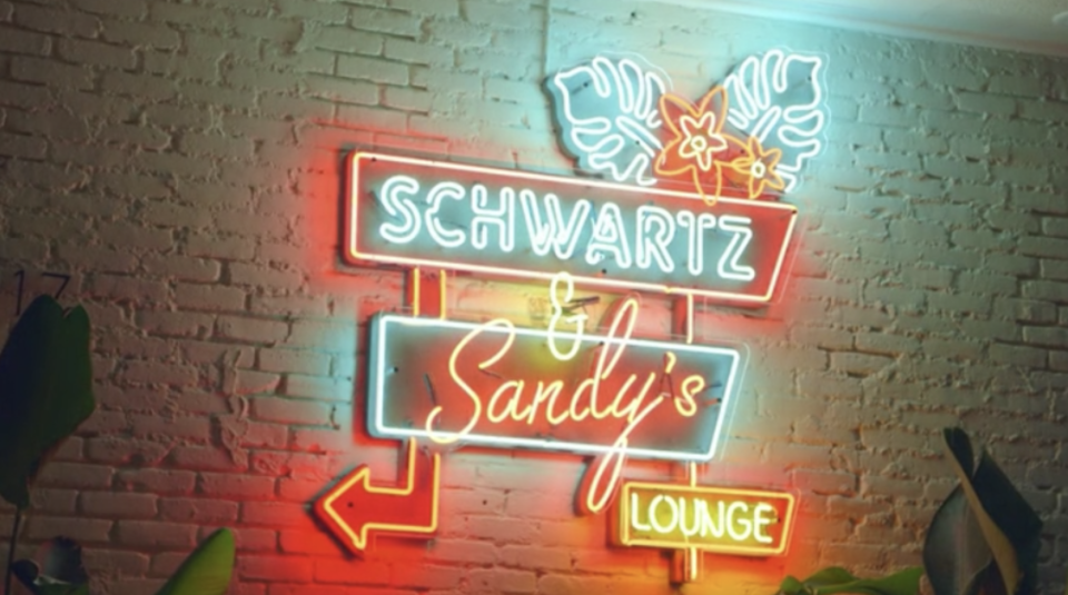 An orange and teal neon sign mounted on a white brick wall, it reads Schwartz & Sandy's Lounge