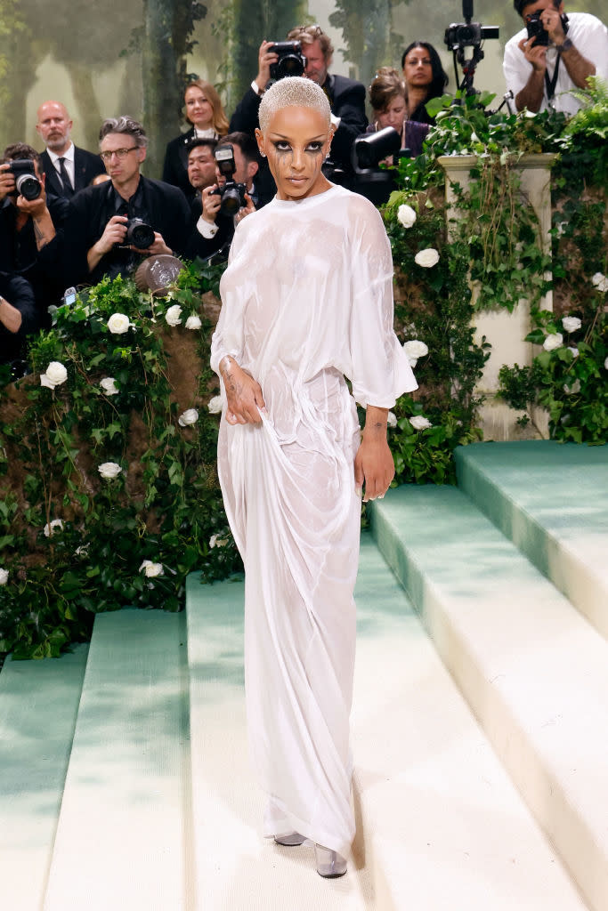 Person in a sheer, draped outfit posing on steps, photographers in background