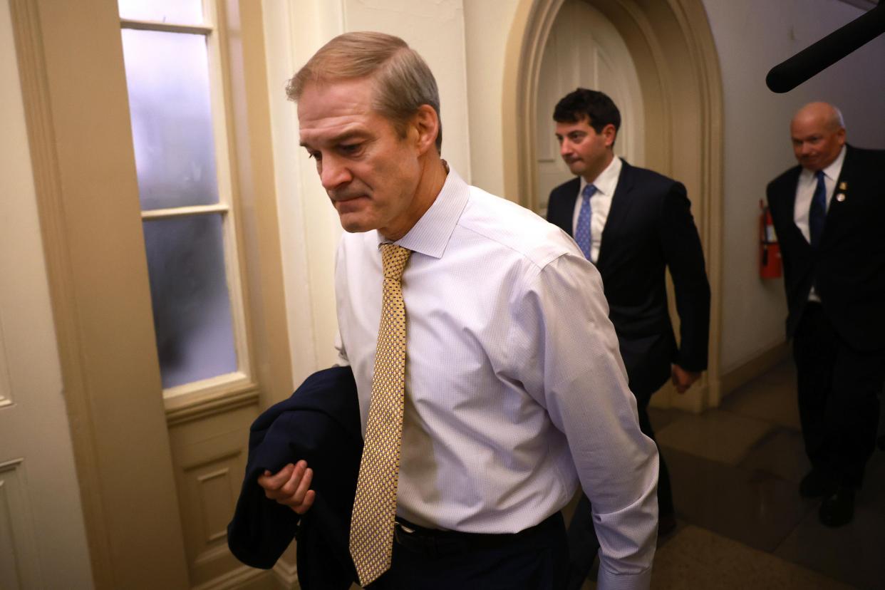 A middle-aged white man wearing a light-blue shirt and yellow tie looks down as he walks, holding his blazer across his arm.