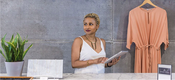 A merchant holding a tablet with a dress that she is selling on a hanger behind her.