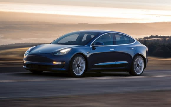 Dark-colored Tesla Model 3 vehicle on a road in front of an open landscape of rolling hills and layered clouds.