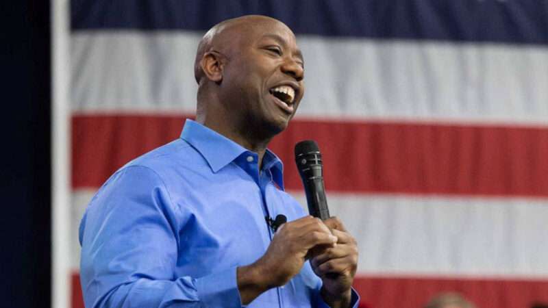Tim Scott standing in front of an American flag, holding a microphone, and looking optimistic.
